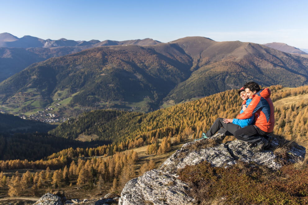 Paar sitzt auf Felsen mit weitem Blick über herbstliche Berge und Täler.