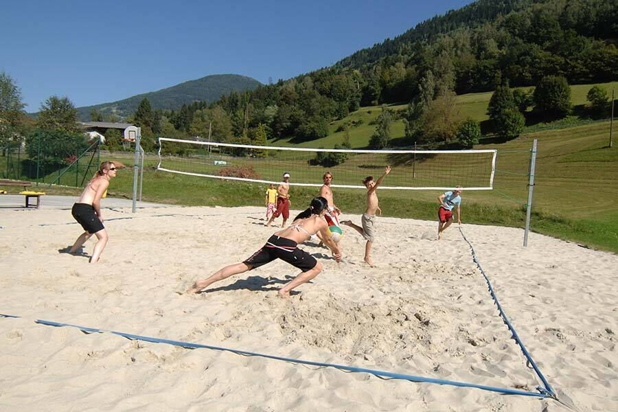 People playing beach volleyball on a sand court in a picturesque mountain landscape.