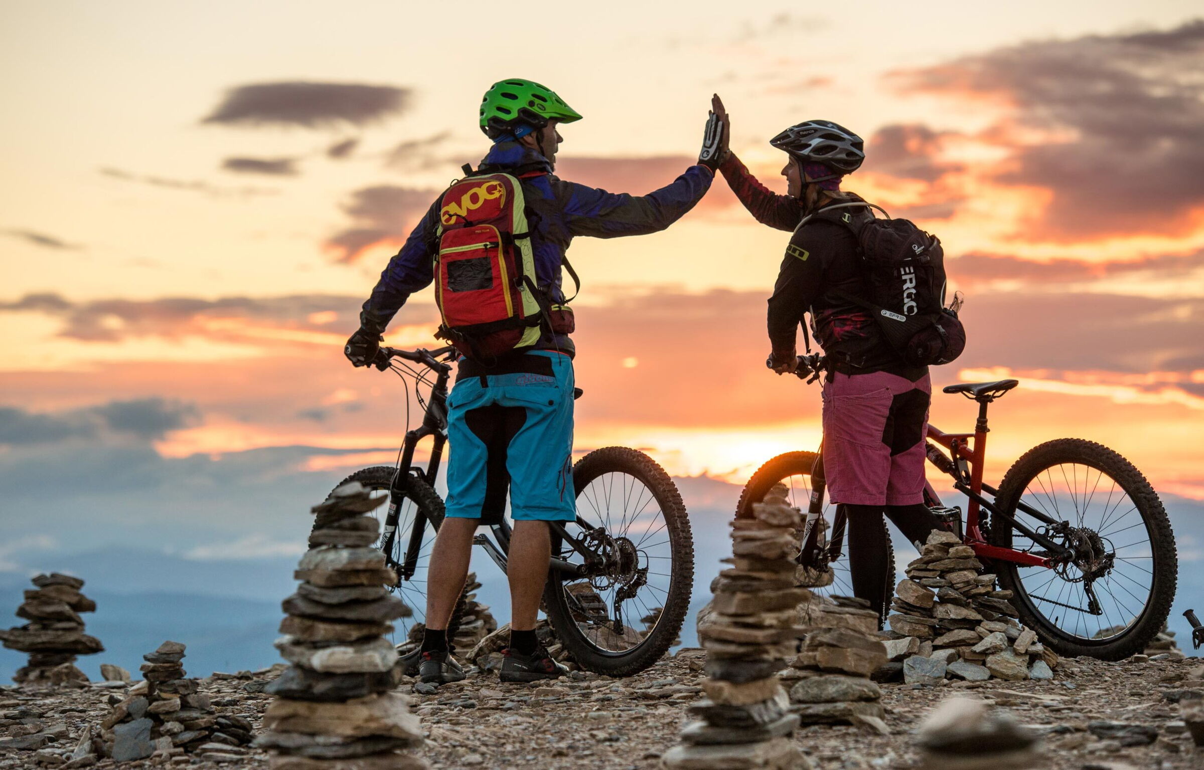 Zwei Mountainbiker genießen den Sonnenuntergang auf einem Gipfel mit Steinmännchen.