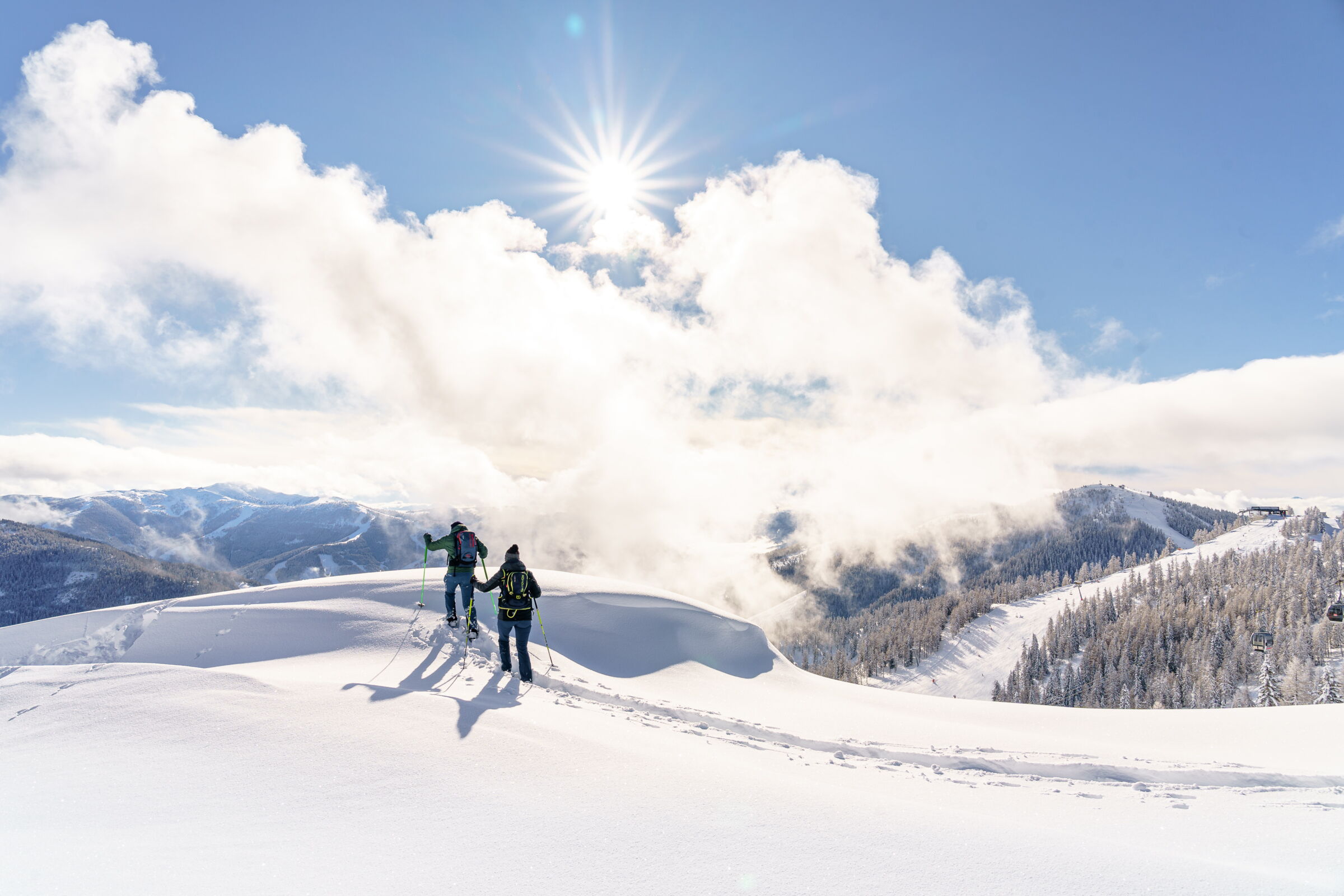 Zwei Personen wandern mit Schneeschuhen unter strahlender Sonne in den verschneiten Bergen.