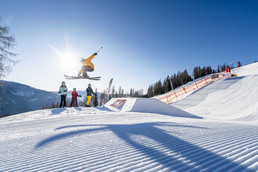 Skifahrer springt über Hügel, Familie schaut zu, blauer Himmel im Hintergrund.