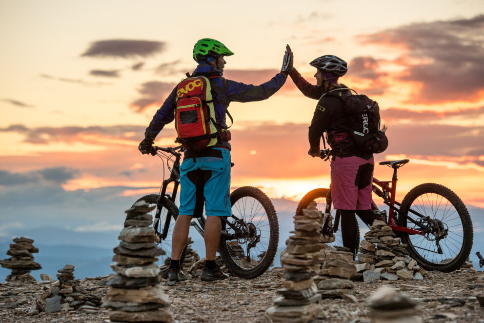 Zwei Mountainbiker geben sich High-Five vor malerischem Sonnenuntergang in den Nockbergen.