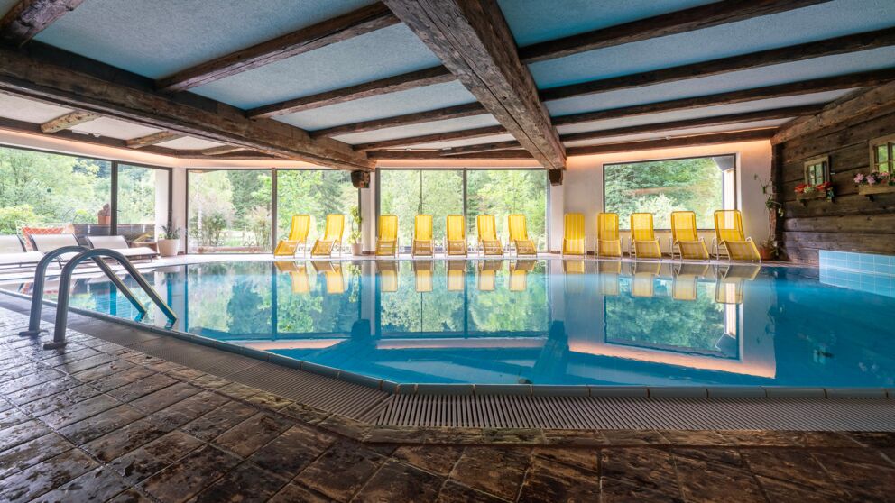 Piscina interna con soffitto in travi di legno e grandi finestre, circondata da lettini gialli.