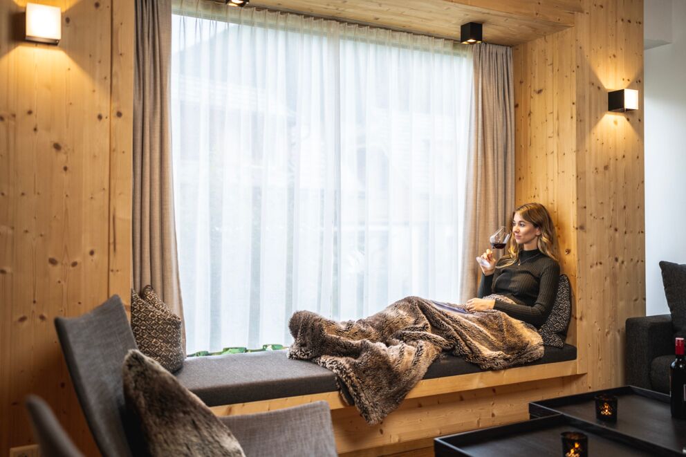Frau genießt Wein auf gemütlicher Sitzbank im Chalet mit weichem Fell und Kerzenlicht.
