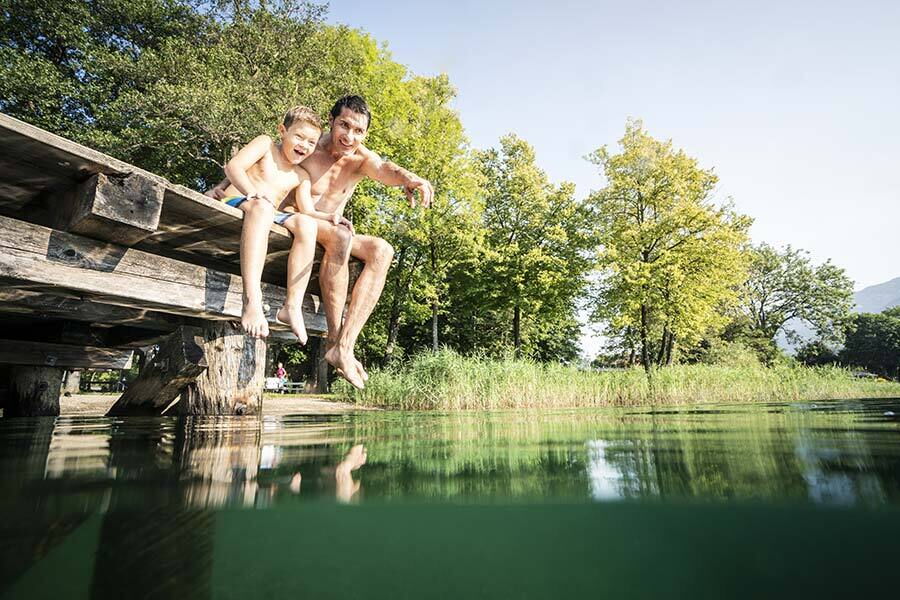 Father and son sit laughing on a jetty above clear water.