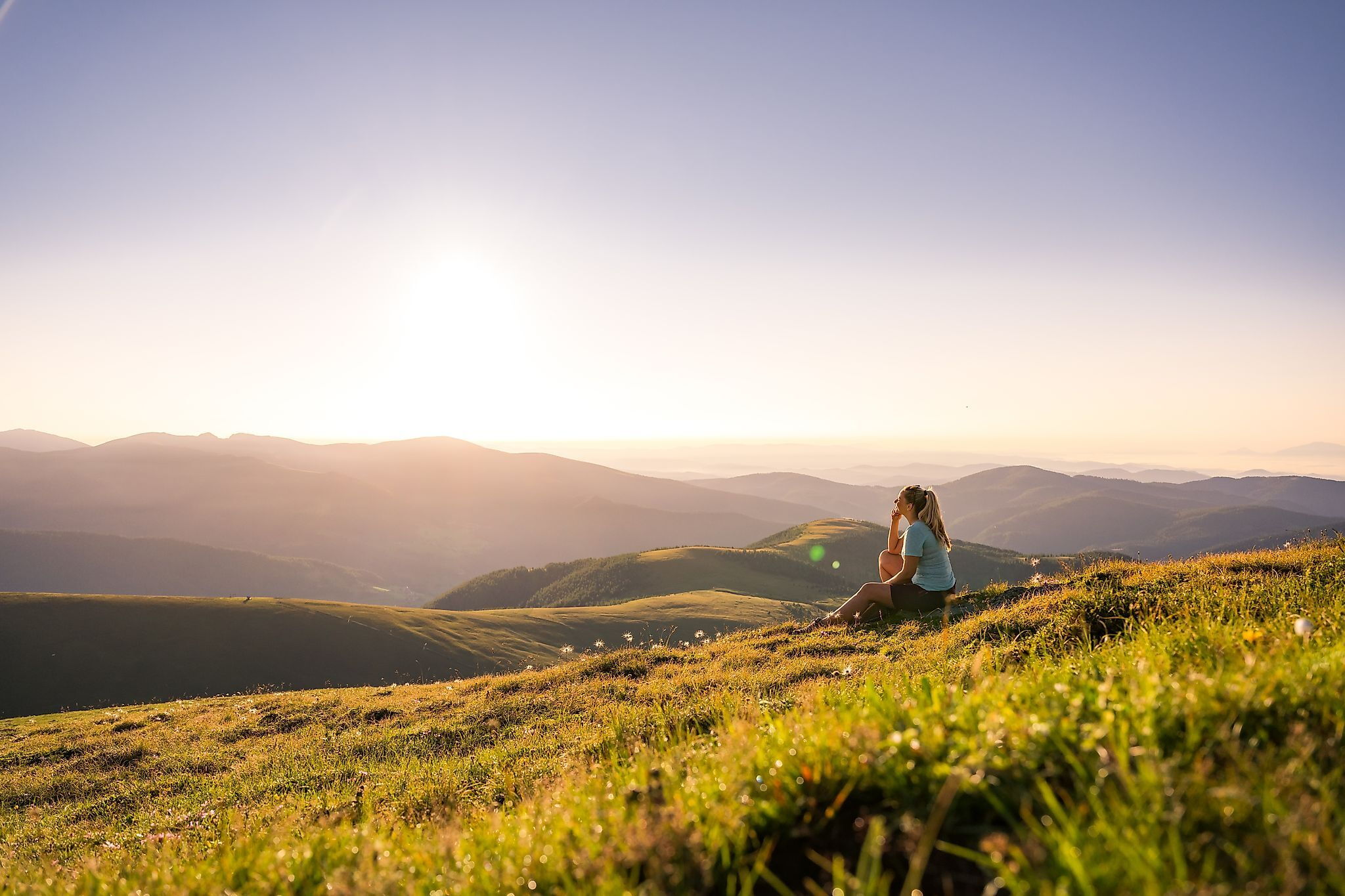 Frau sitzt auf Wiese mit weitem Blick über sonnige Berglandschaft.