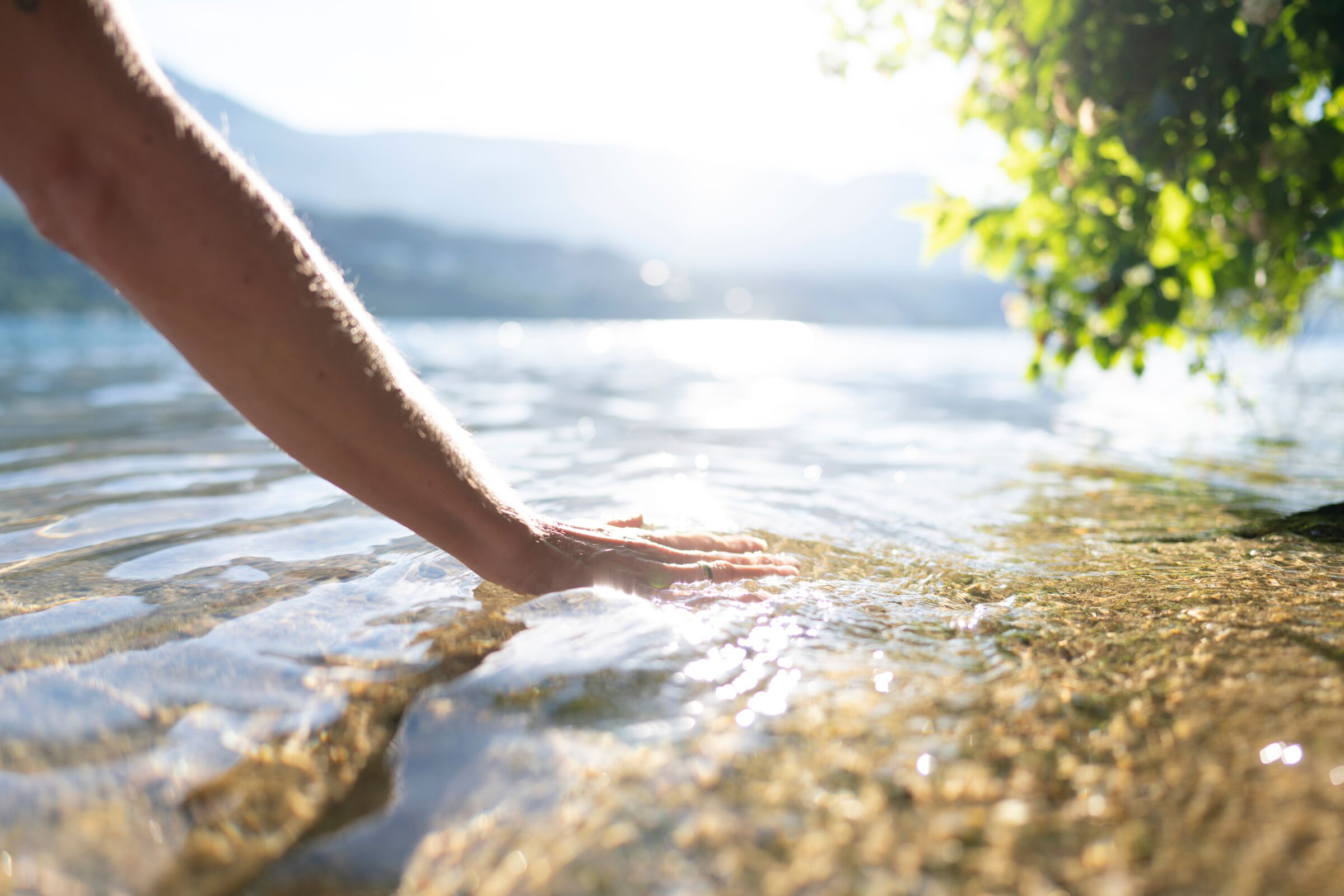 Hand gently touches the clear water on the lakeshore, surrounded by sunlight