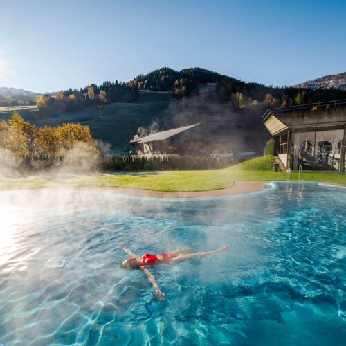 Person relaxes in the steaming outdoor pool with a view of picturesque mountains and blue skies.