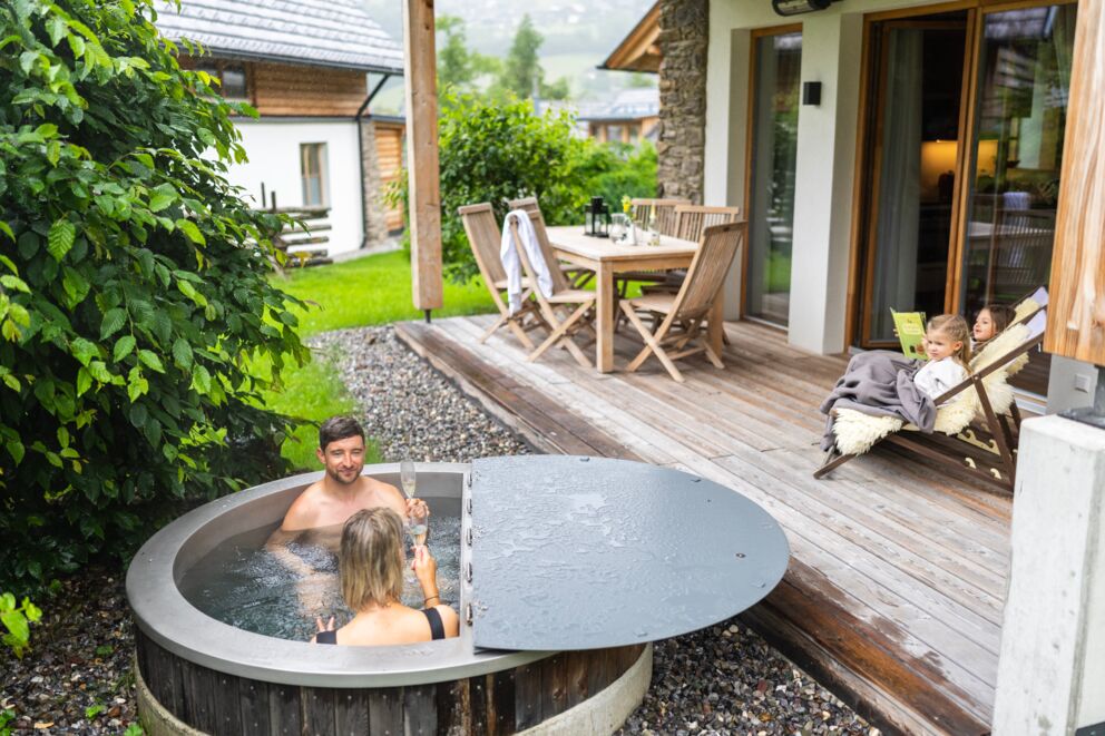 Zwei Erwachsene im Whirlpool, Kinder im Liegestuhl, gemütliches Chalet im Hintergrund.