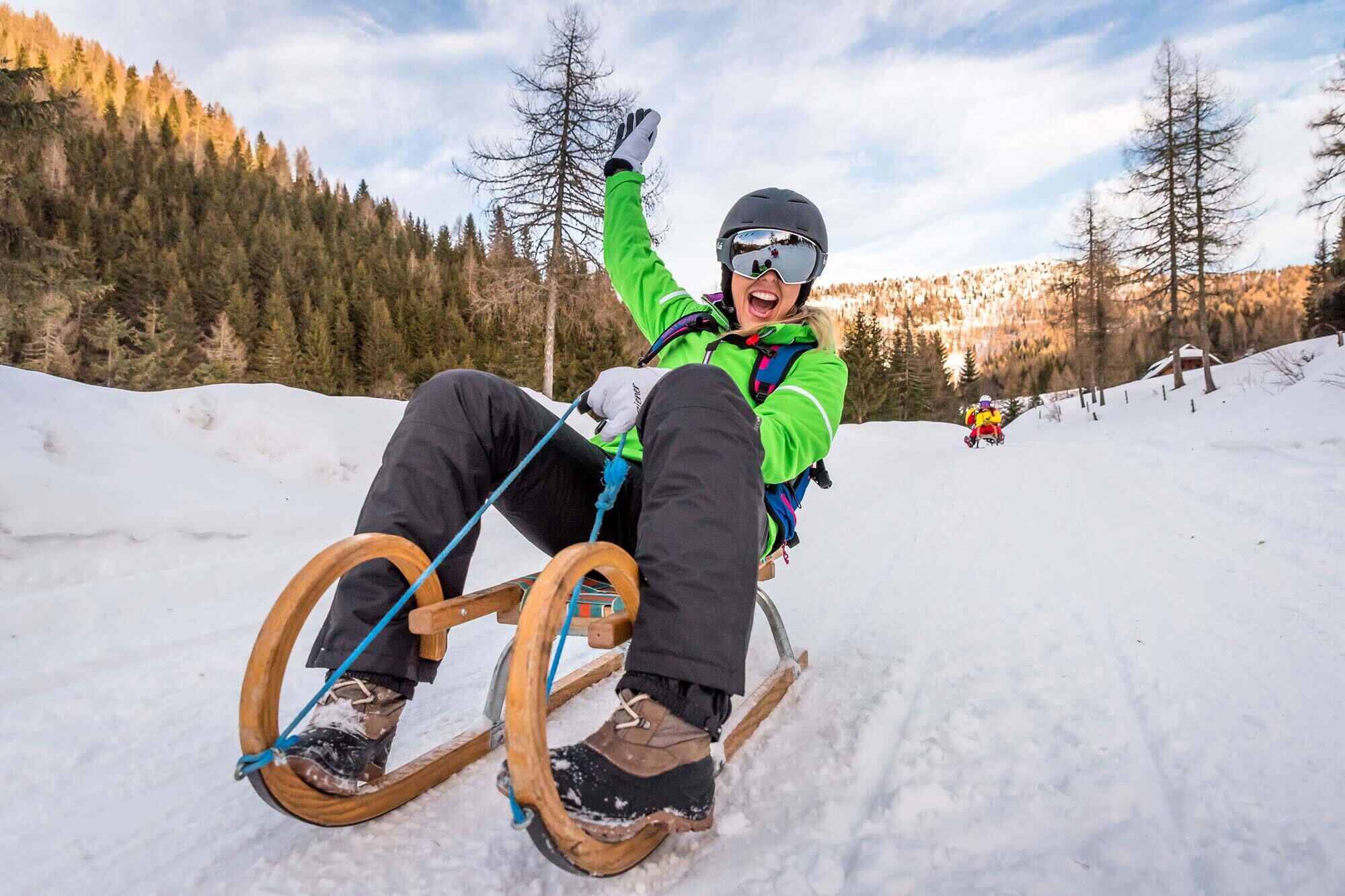 Person auf Schlitten im Schnee, umgeben von verschneiten Bäumen und Bergen.