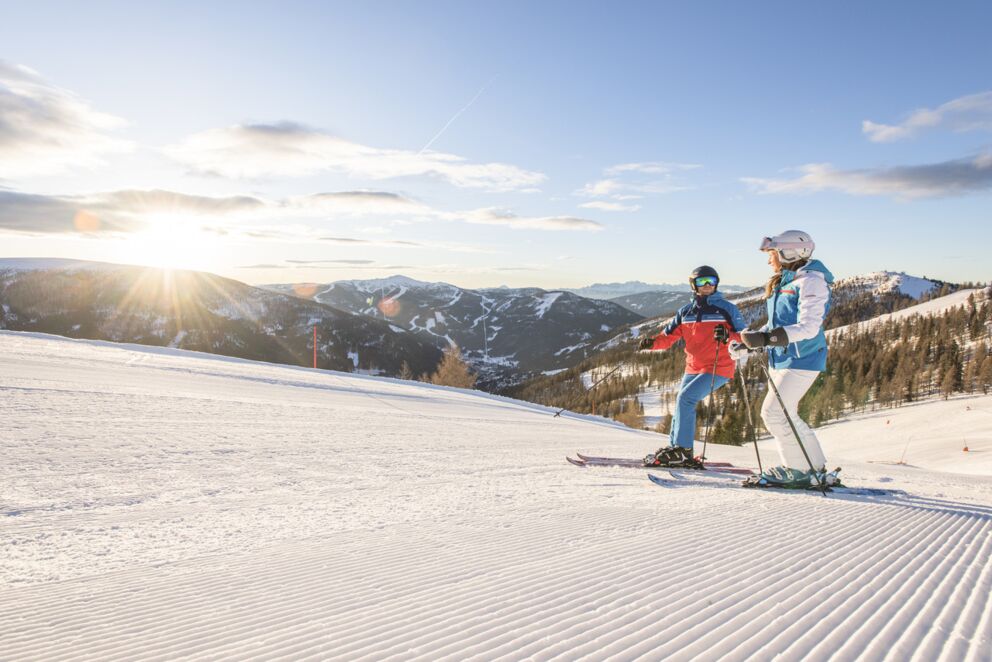Zwei Skifahrer auf frisch präparierter Piste mit strahlendem Sonnenschein im Hintergrund.