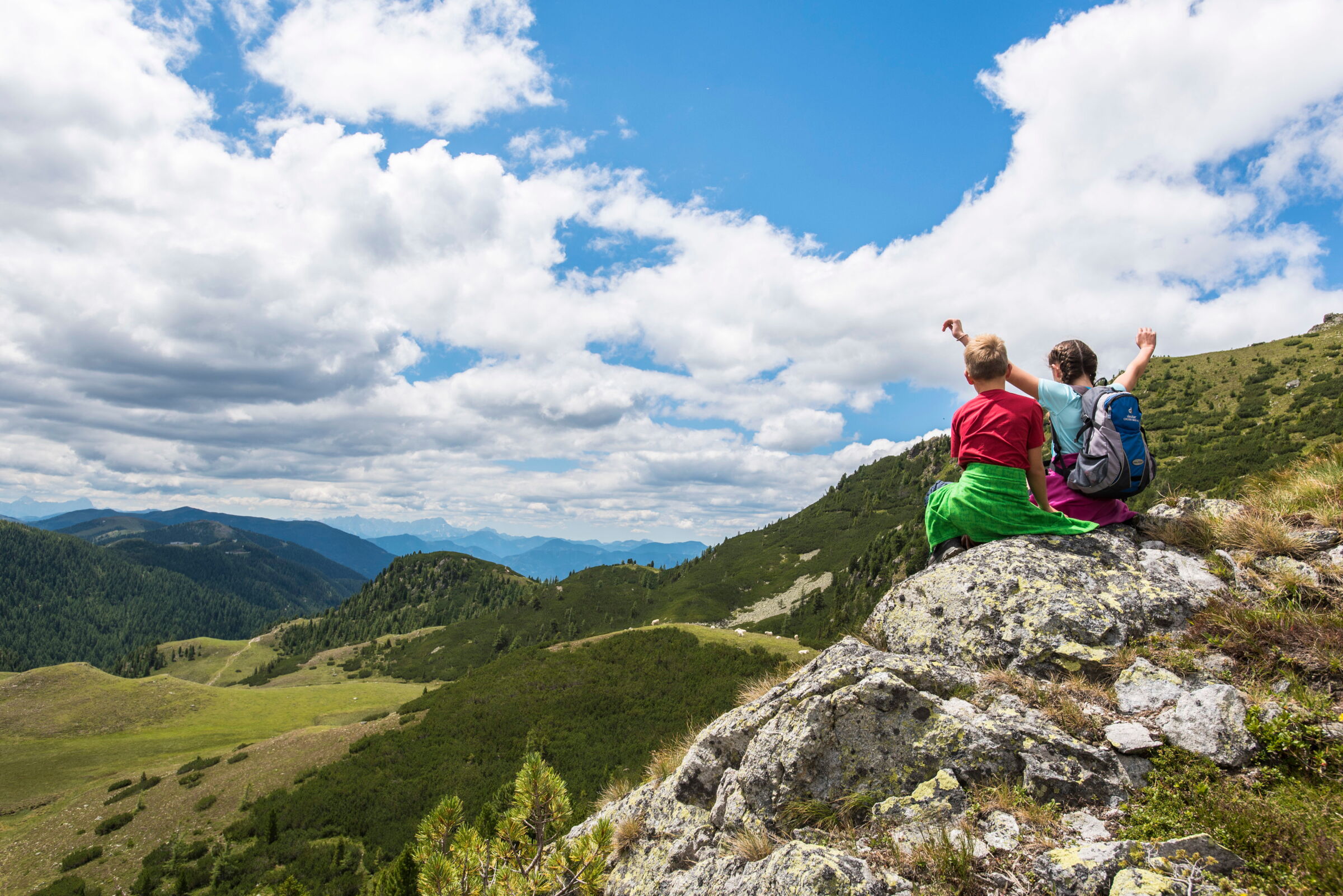 Zwei Kinder sitzen auf einem Felsen mit Blick über die Nockberge unter blauem Himmel.