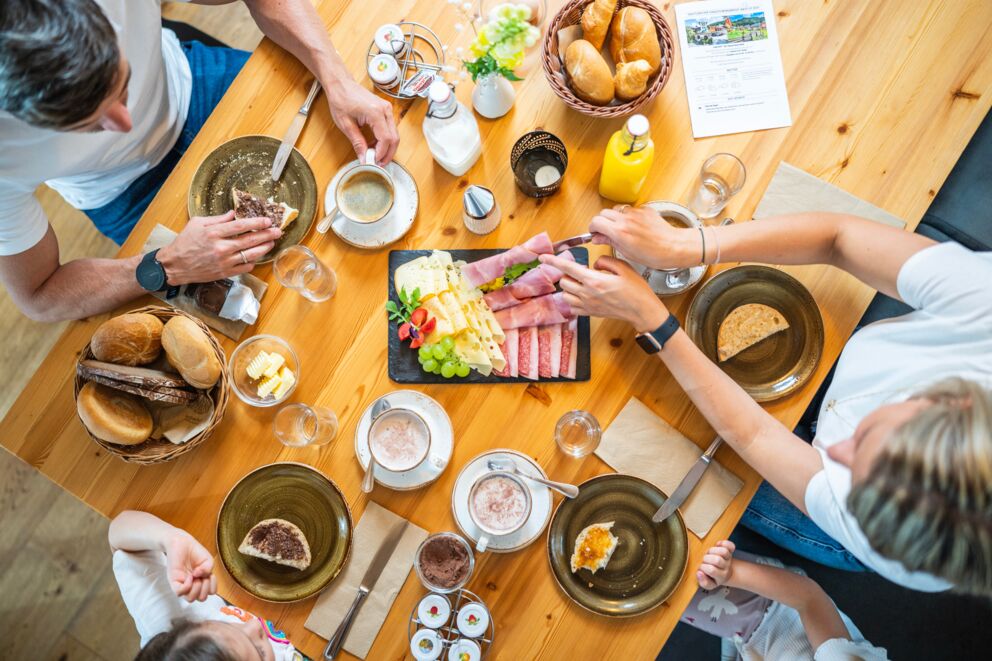 Familienfrühstück mit Aufschnitt, Käse, Brötchen und Getränken auf rustikalem Holztisch.