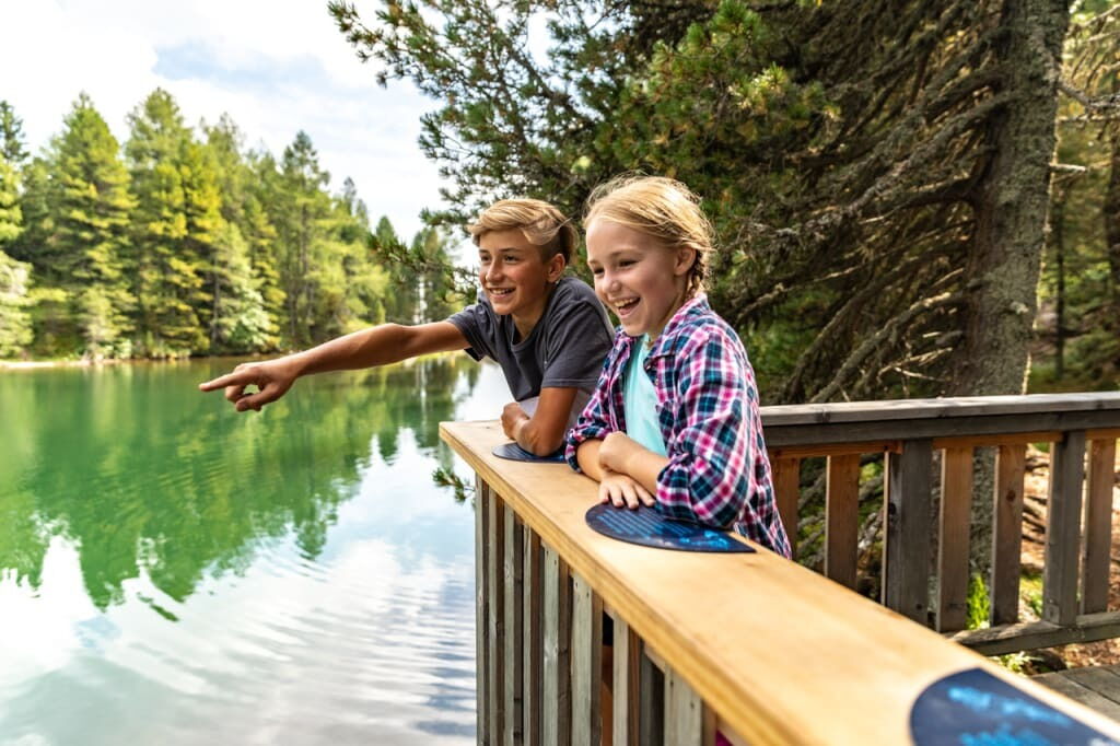 Two laughing children lean against a railing by a peaceful lake in the countryside.