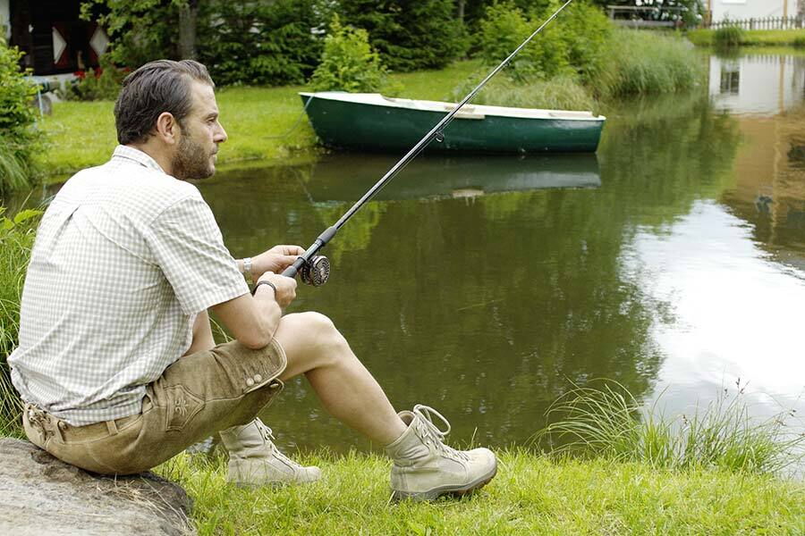 Man fishing relaxed at a fish pond, a boat and lush greenery in the background.