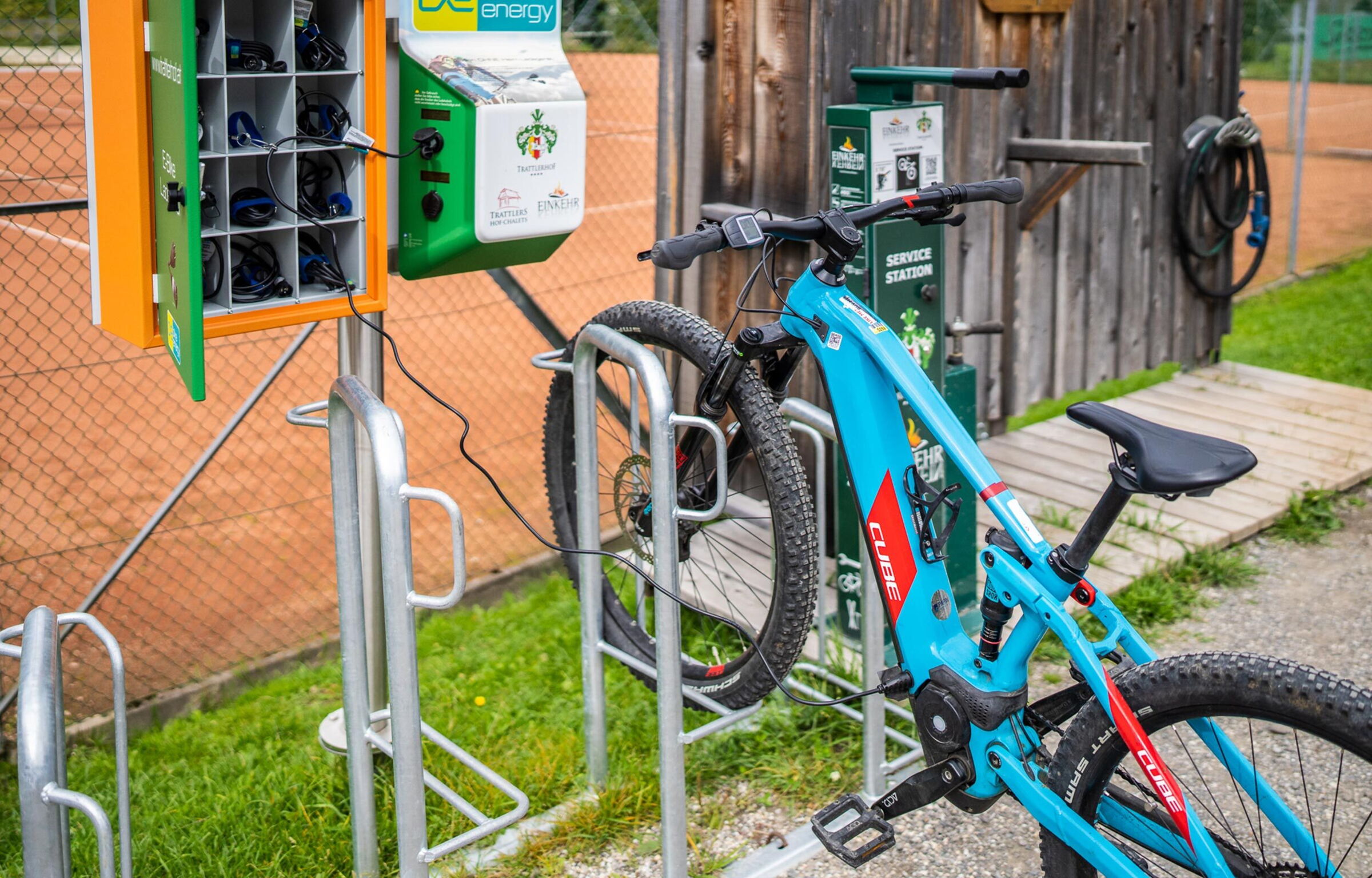 E-bike parcheggiata alla stazione di ricarica accanto a un edificio in legno e a un campo da tennis in un ambiente naturale.