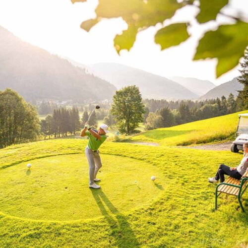Golfer swings a club at sunset, with a mountain panorama in the background.