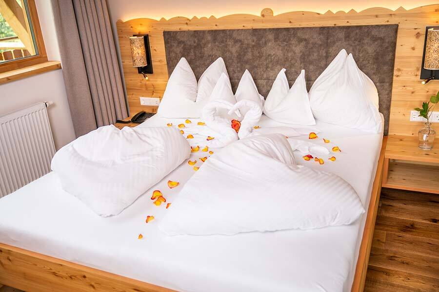 Gemütliches Bett mit dekorativen Kissen und Rosenblättern auf weißer Bettwäsche.