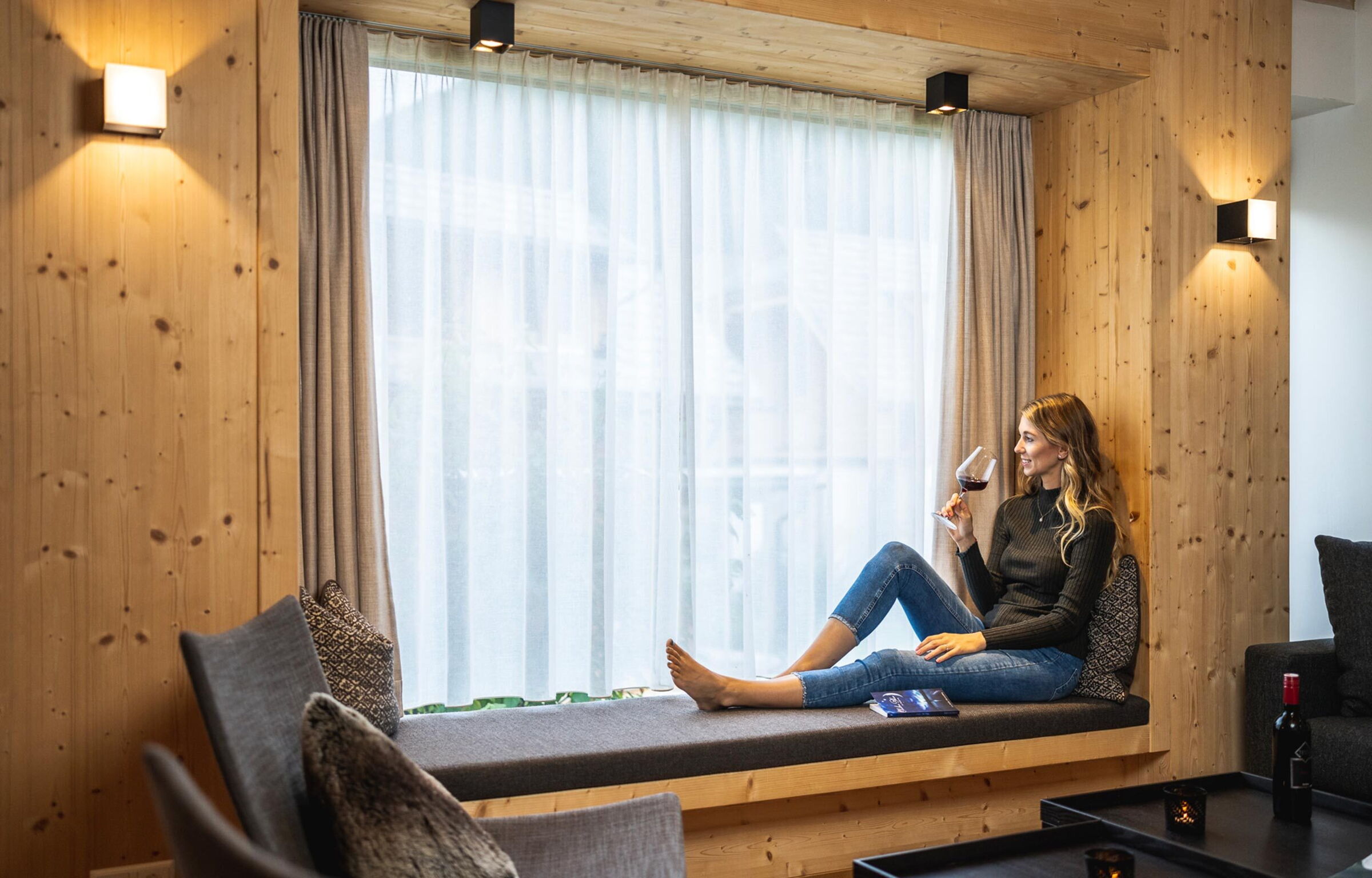 Frau genießt Wein, sitzt gemütlich am Fenster in einem modernen Chalet.