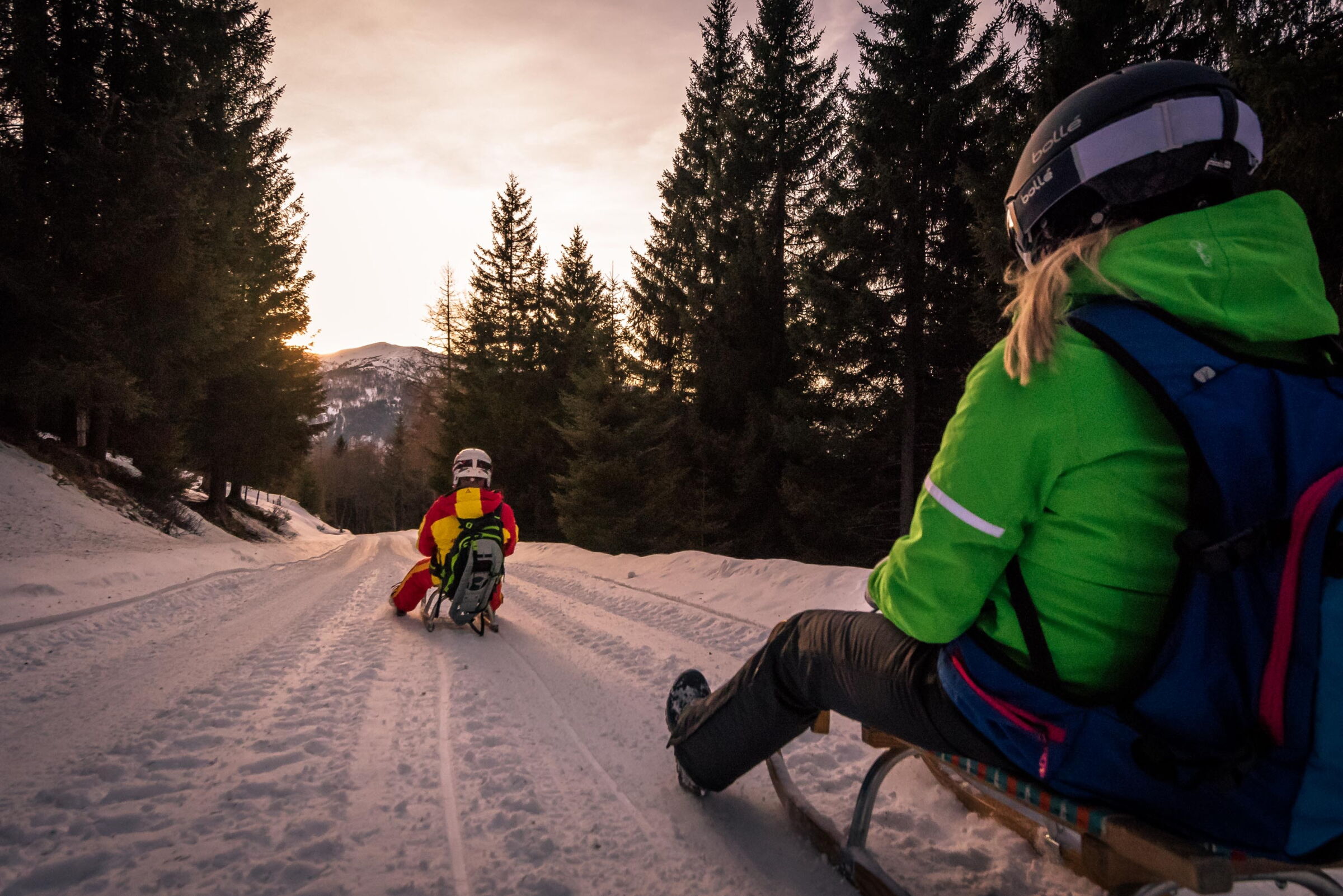 Two people tobogganing at sunset on a snow-covered forest road.