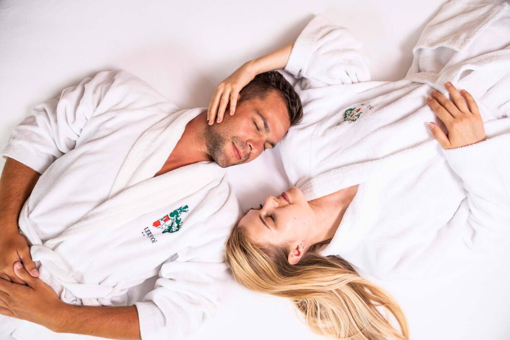 Couple in white bathrobes, lying relaxed on a bed, smiling gently.