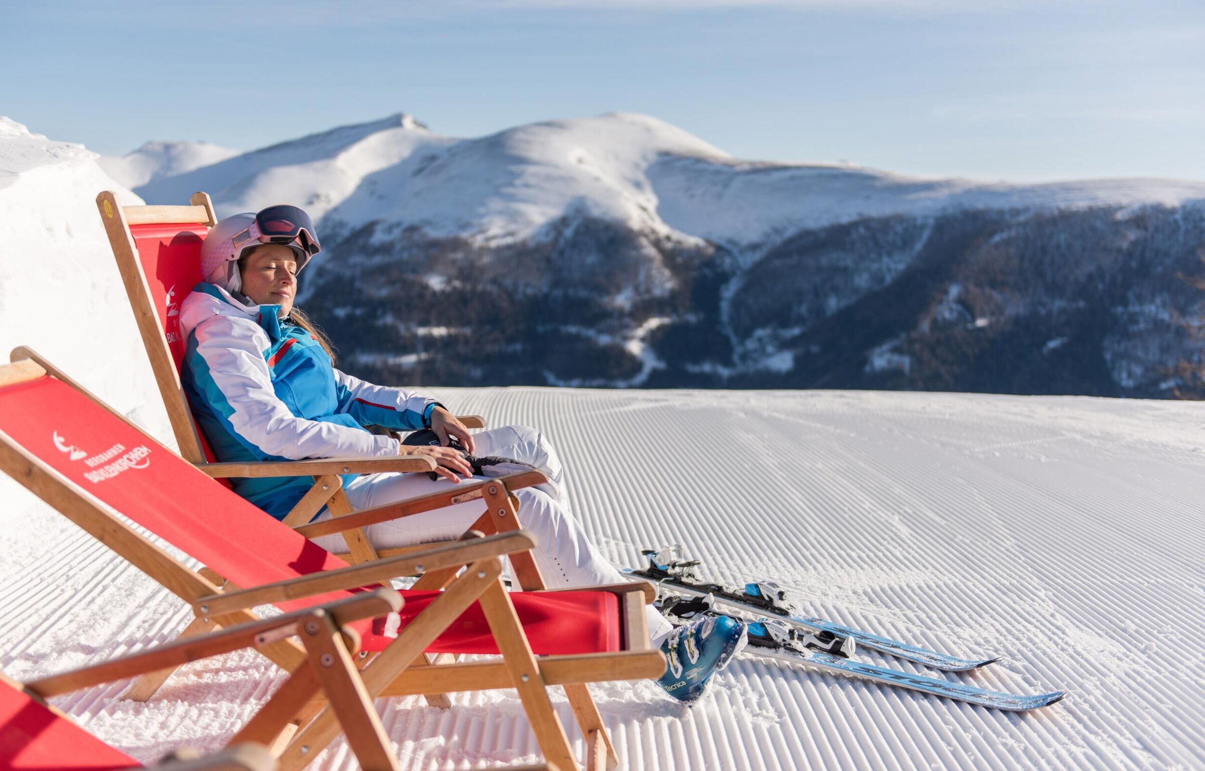 Entspannte Skifahrerin in Sonnenliege mit verschneitem Bergpanorama im Hintergrund.