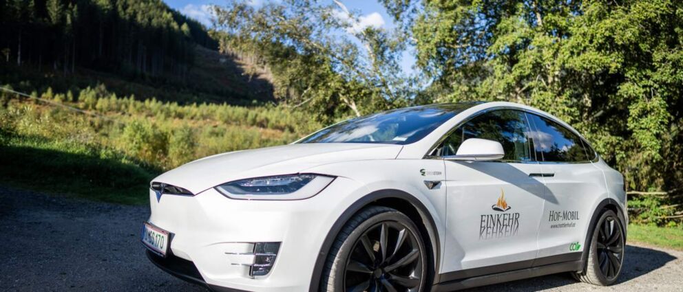 Weißes Elektroauto vor grüner Waldkulisse im Sonnenschein geparkt.