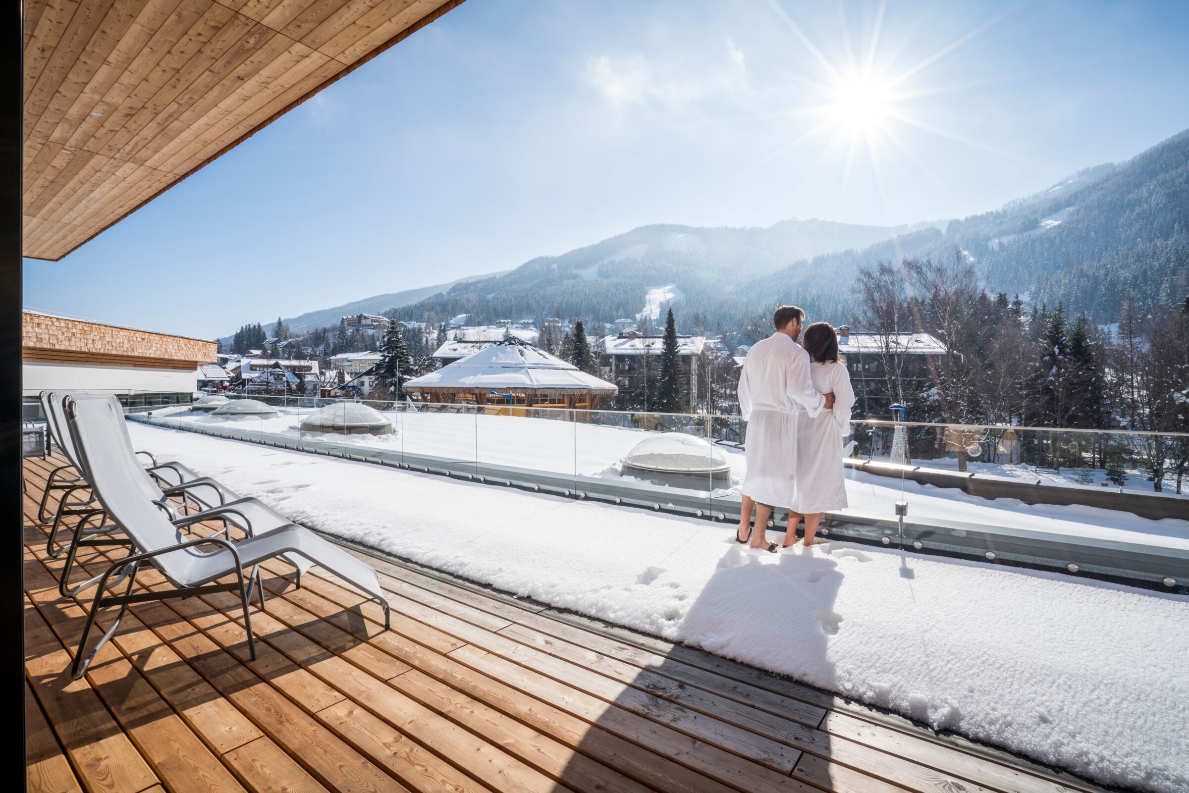 Couple enjoying sunny winter views from a terrace with deckchairs and snow-covered landscape.