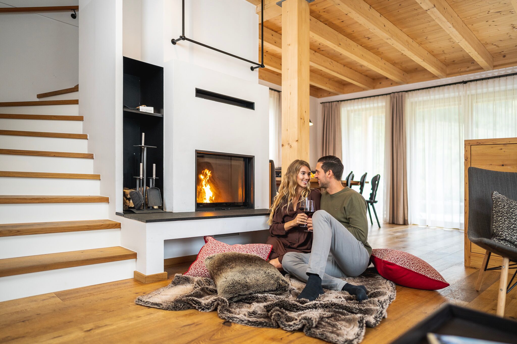 Couple sitting in front of a burning fireplace in a chalet with wooden beams and large windows.
