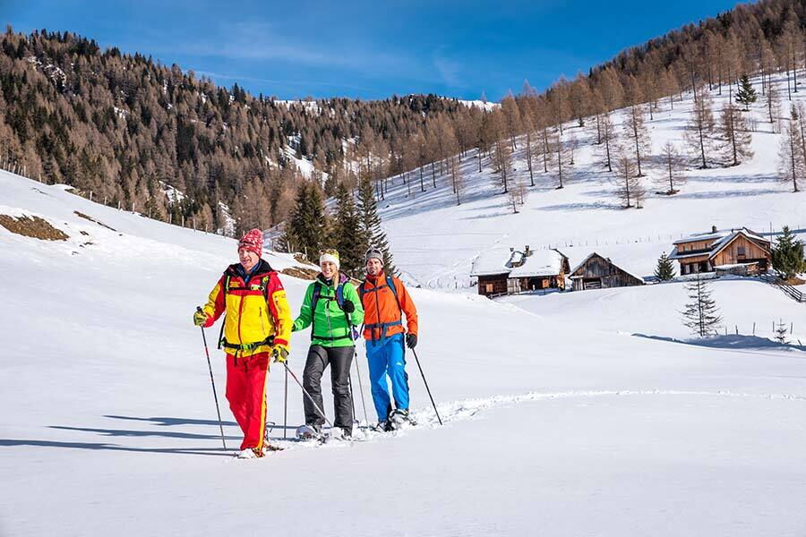 Drei Personen beim Schneeschuhwandern vor schneebedeckten Hütten in den Bergen.