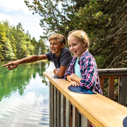 Zwei Kinder lachen und zeigen auf einen idyllischen See im Wald.
