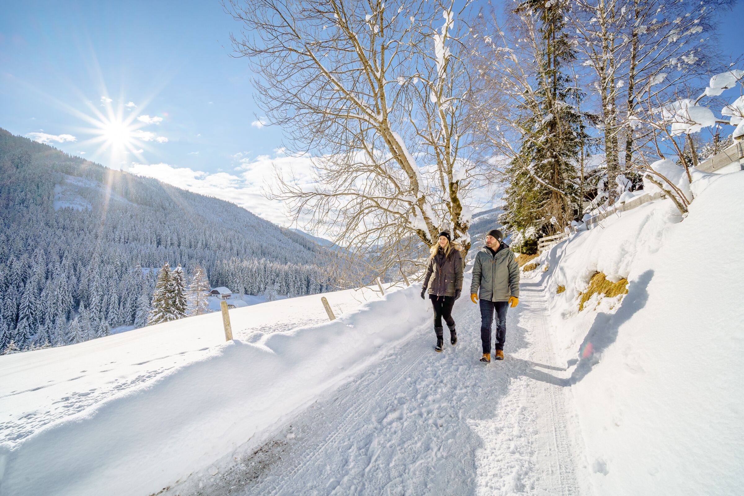 Zwei Personen wandern auf verschneitem Pfad in winterlicher Berglandschaft bei Sonnenschein.