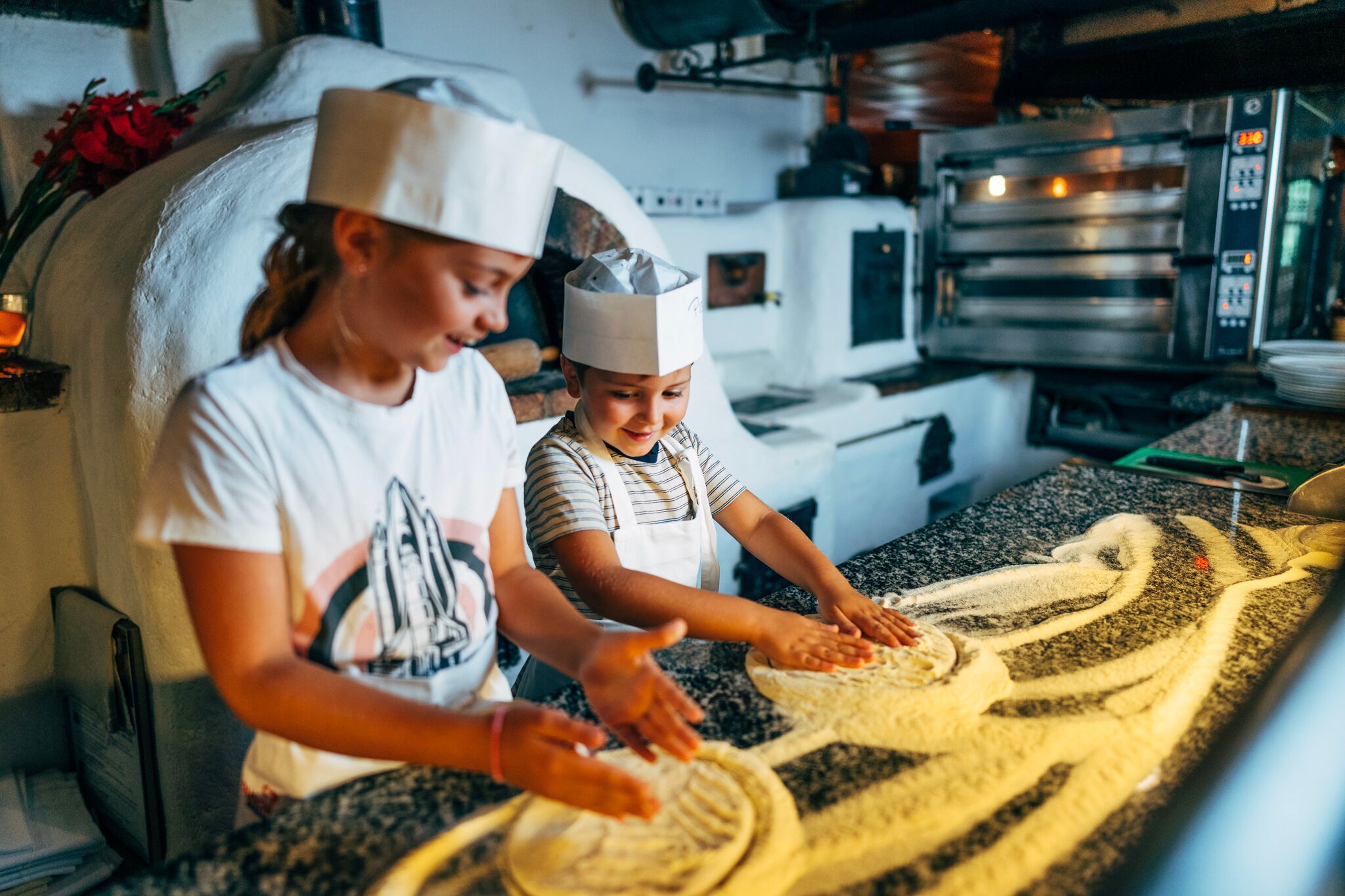 Two children with chef's hats knead dough on a floured work surface.