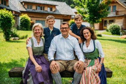 Four adults and two children in traditional dress in front of wooden houses in the countryside.