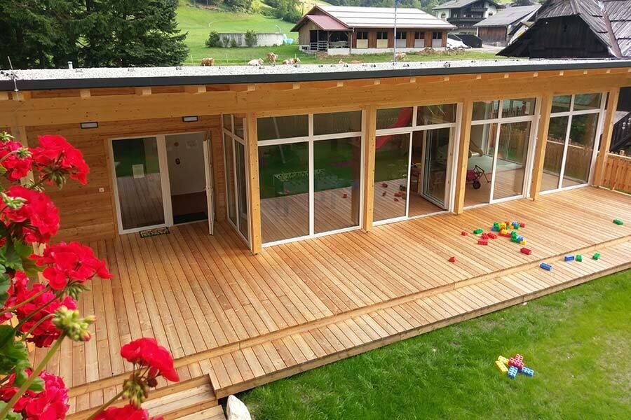 Veranda in legno con vetrata, area giochi e prato verde circondato da fiori.