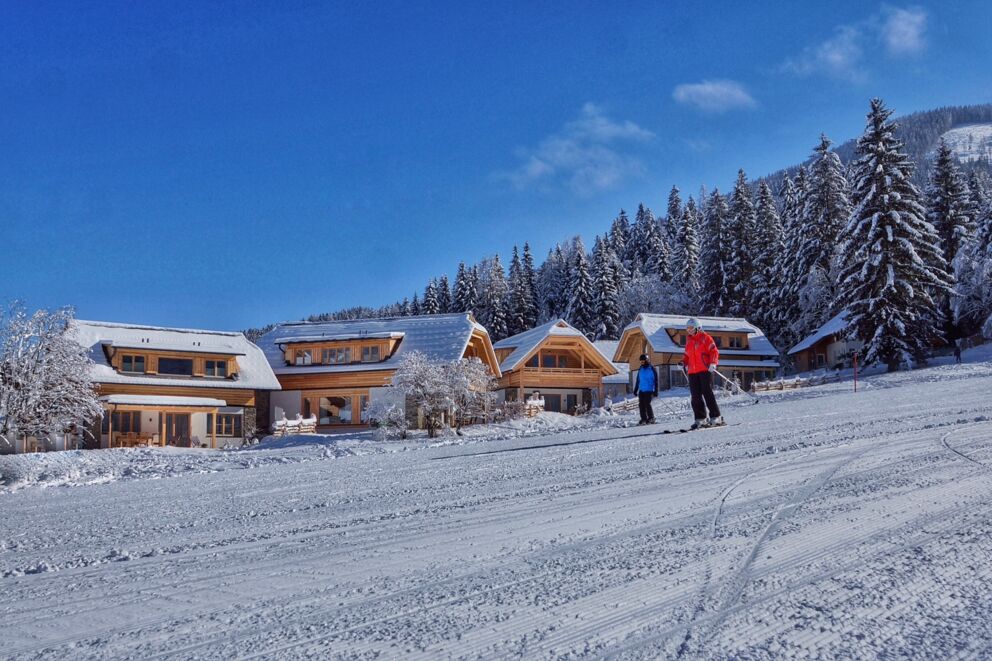 Verschneite Chalets neben Skipiste, umgeben von schneebedeckten Bäumen und strahlend blauem Himmel.