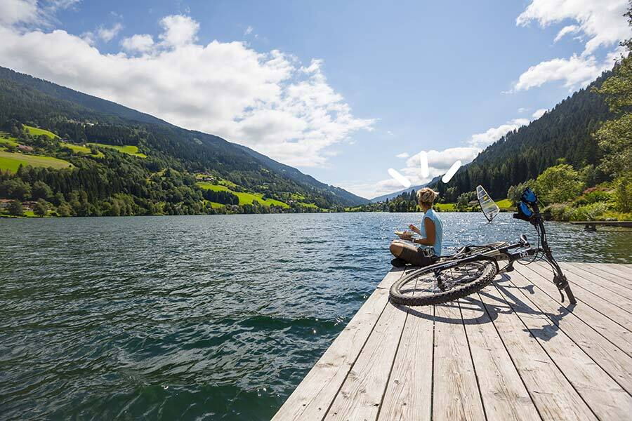 Person sits with bicycle on jetty in front of a picturesque lake and mountains.