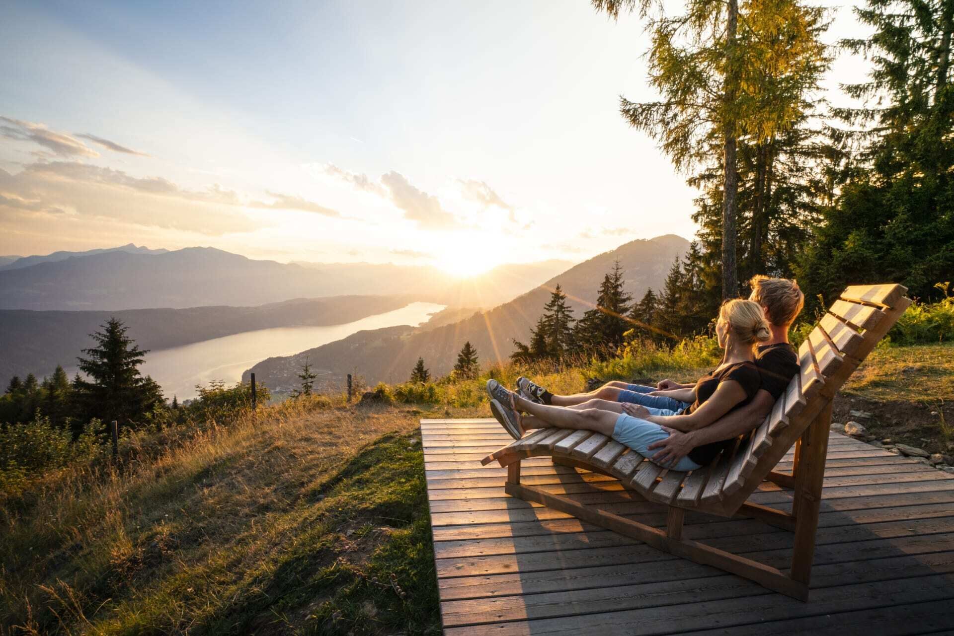 Couple relaxing on a wooden lounger with a view of the lake at sunset.