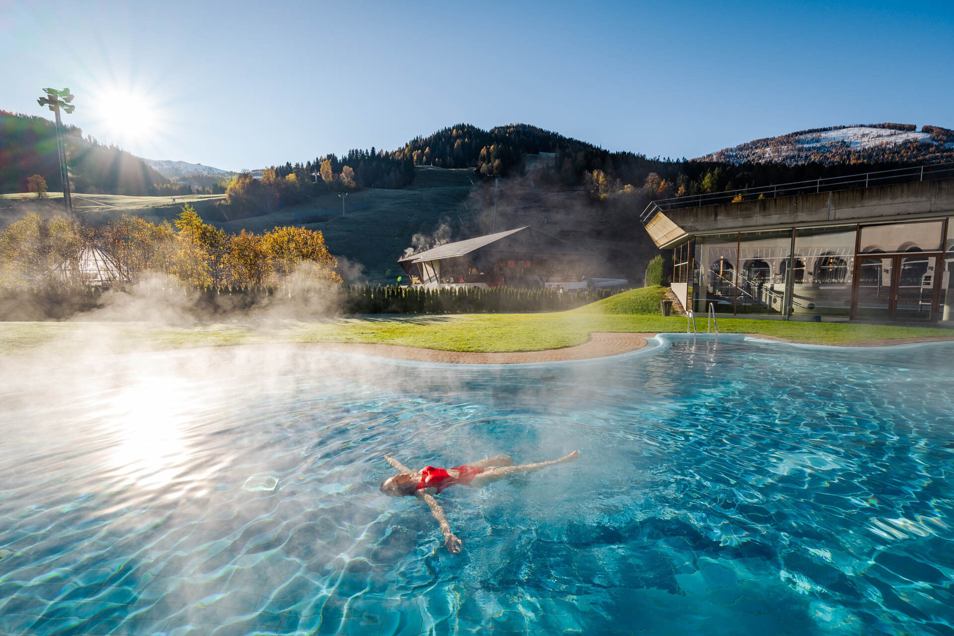 Person entspannt im dampfenden Außenpool mit Blick auf malerische Berge und blauen Himmel.