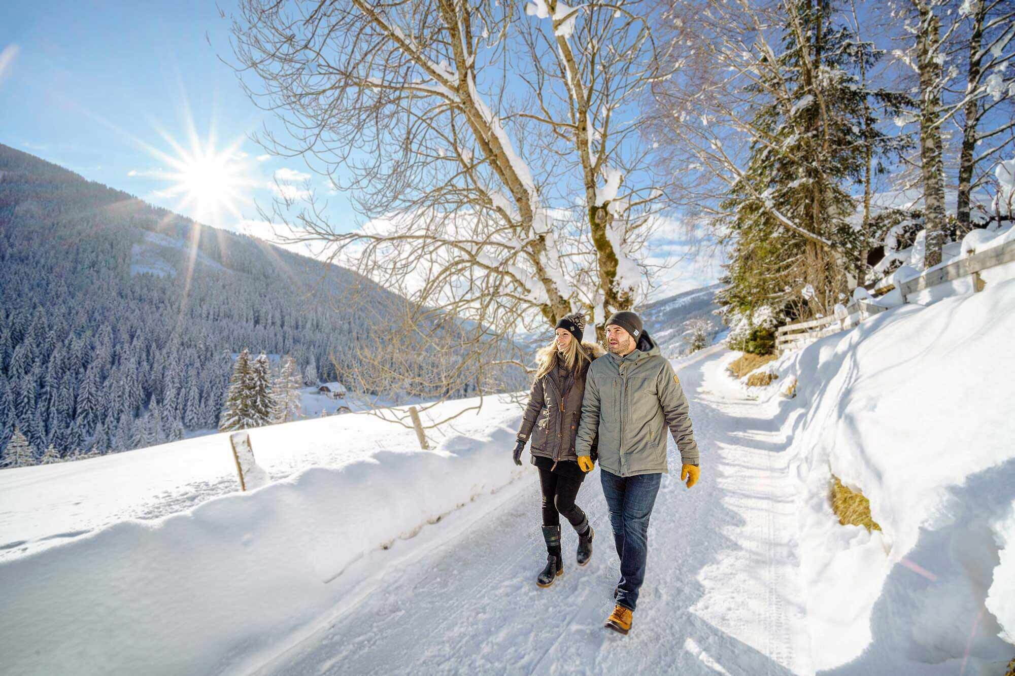 Paar wandert auf verschneitem Pfad bei strahlendem Sonnenschein durch eine winterliche Berglandschaft.