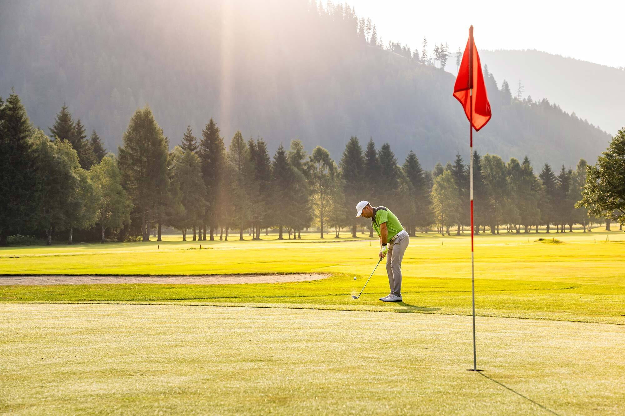 Golfer beim Abschlag auf grünem Platz, umgeben von Bäumen und sanftem Sonnenlicht.