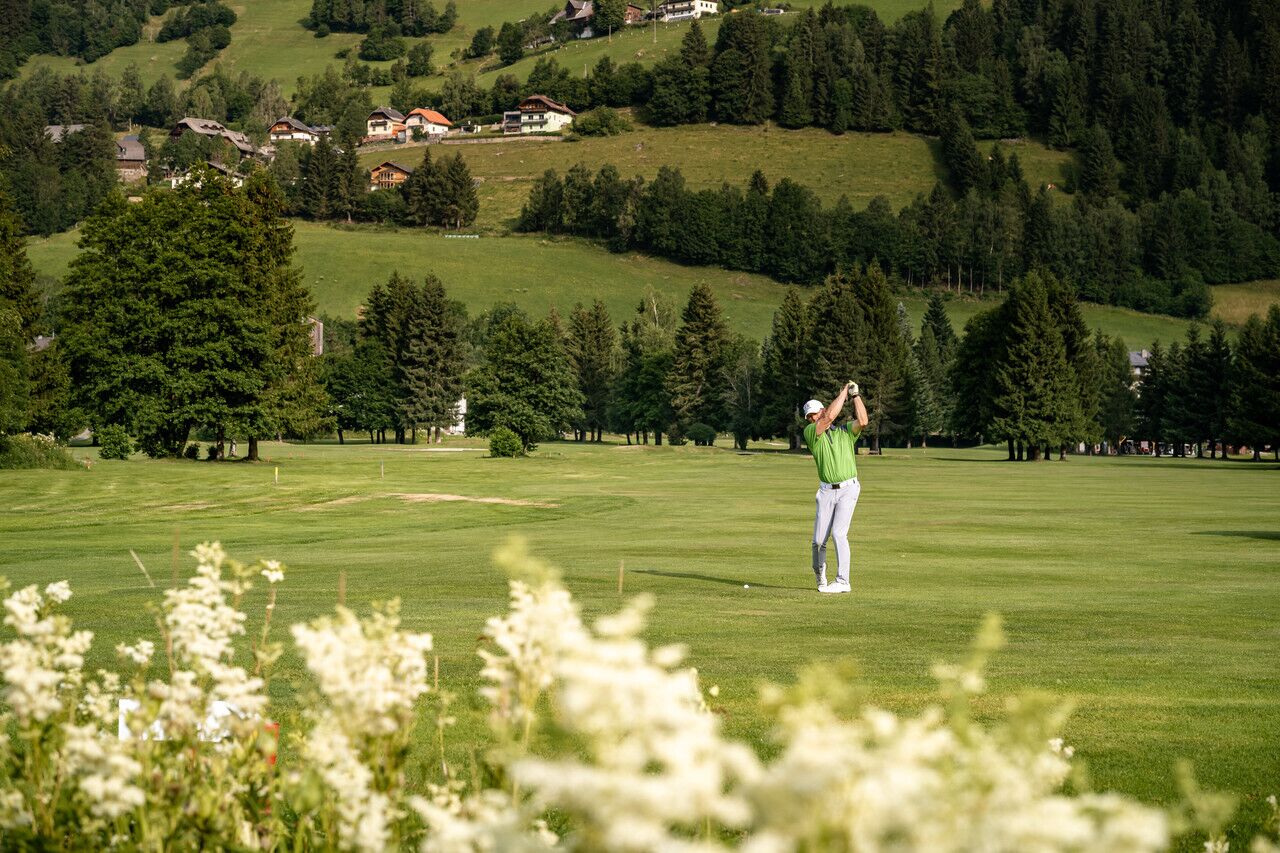 Giocatore di golf colpisce una palla su un green curato con un paesaggio montano sullo sfondo.