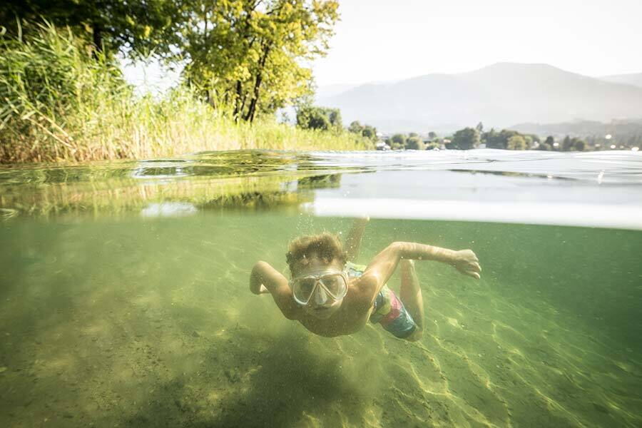 A child with a diving mask swims in a clear lake surrounded by green trees.