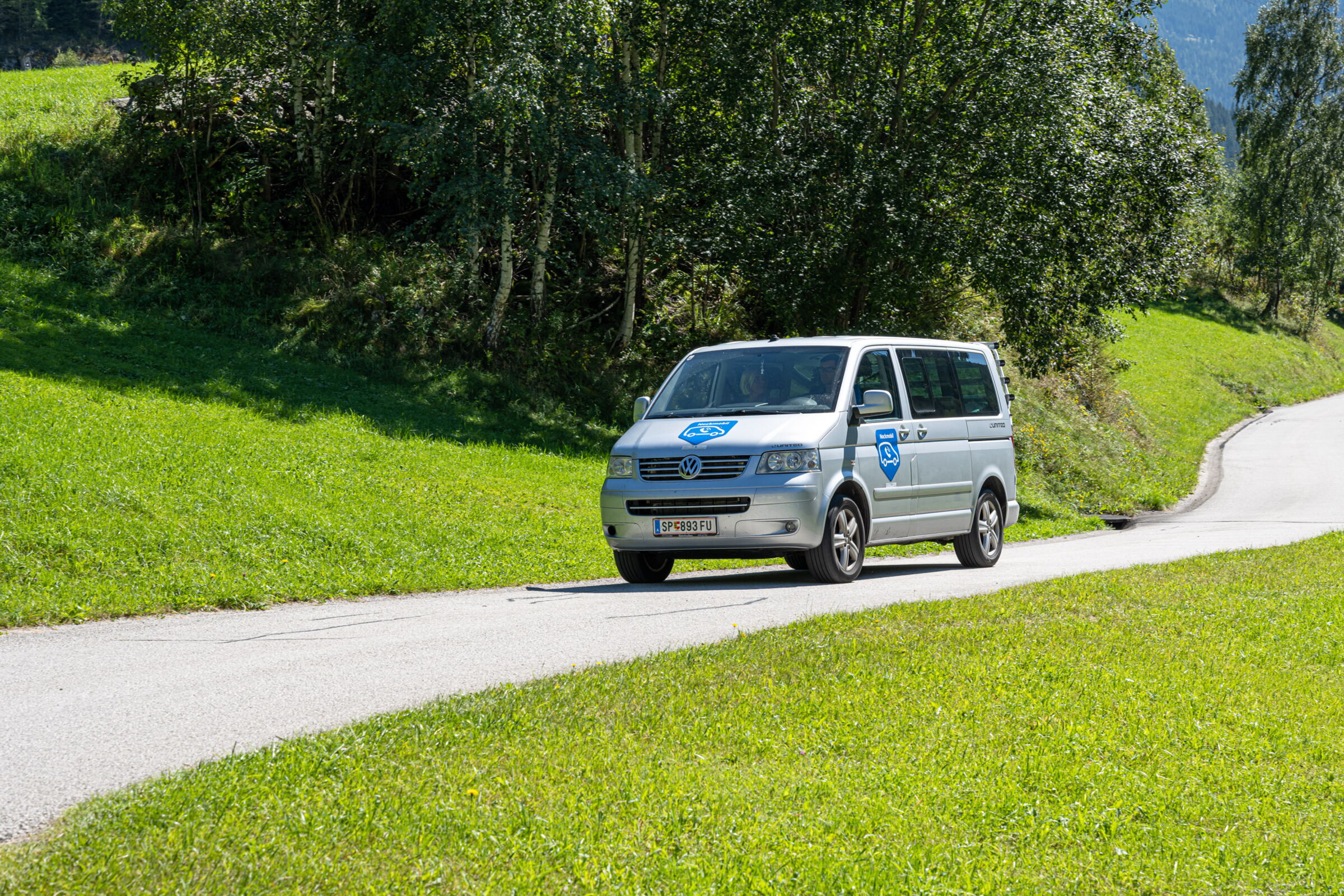 Silberner Shuttle-Bus auf sonniger Landstraße, umgeben von grünen Wiesen.