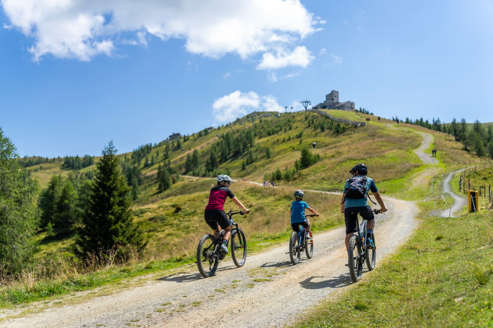 Familie radelt auf sonnigem Bergpfad mit klarblauem Himmel und grüner Landschaft.