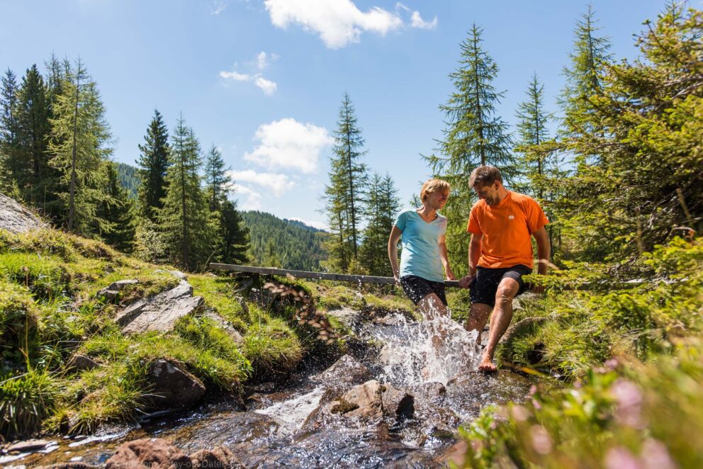 Couple enjoying a sunny hike through a lush mountain landscape with a bubbling stream.