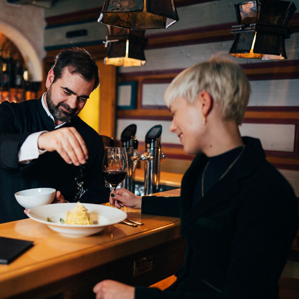 Chef refines dish, woman enjoys red wine in a cozy atmosphere