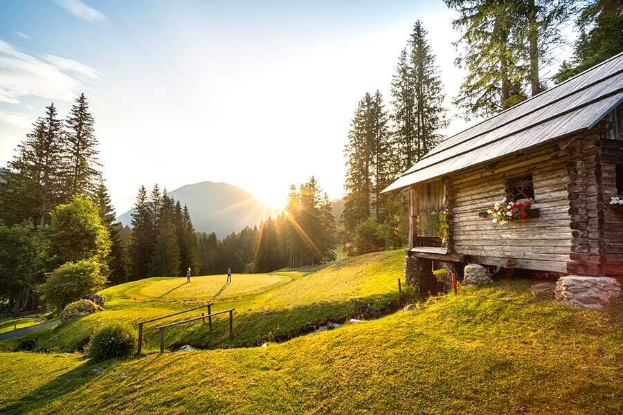 Holzchalet am Waldrand mit Sonnenaufgang über Wiesen und Bäumen.