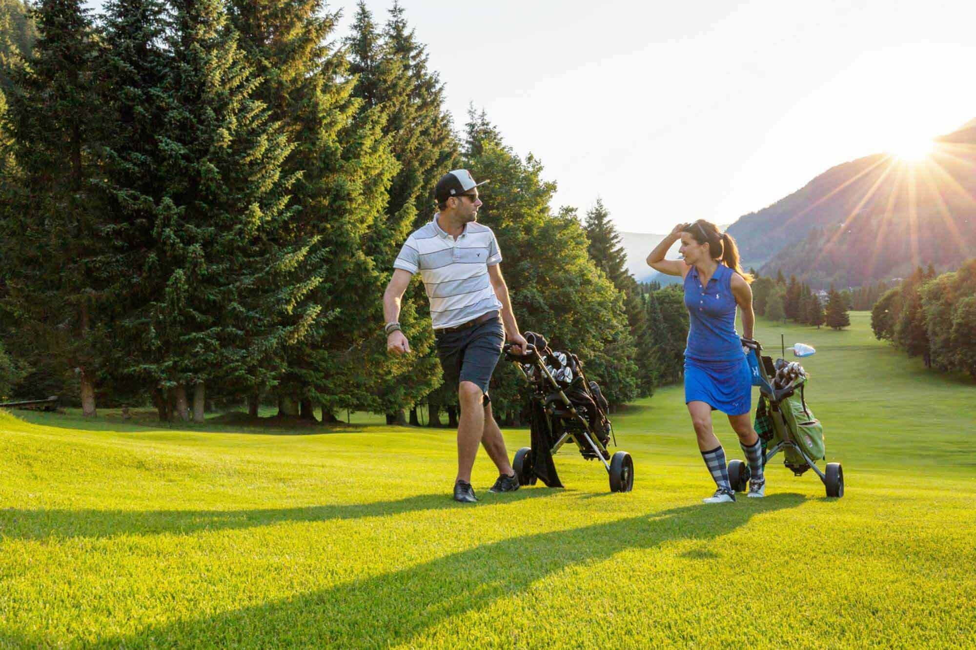 Zwei Personen genießen Golfspiel bei Sonnenaufgang vor malerischer Bergkulisse.