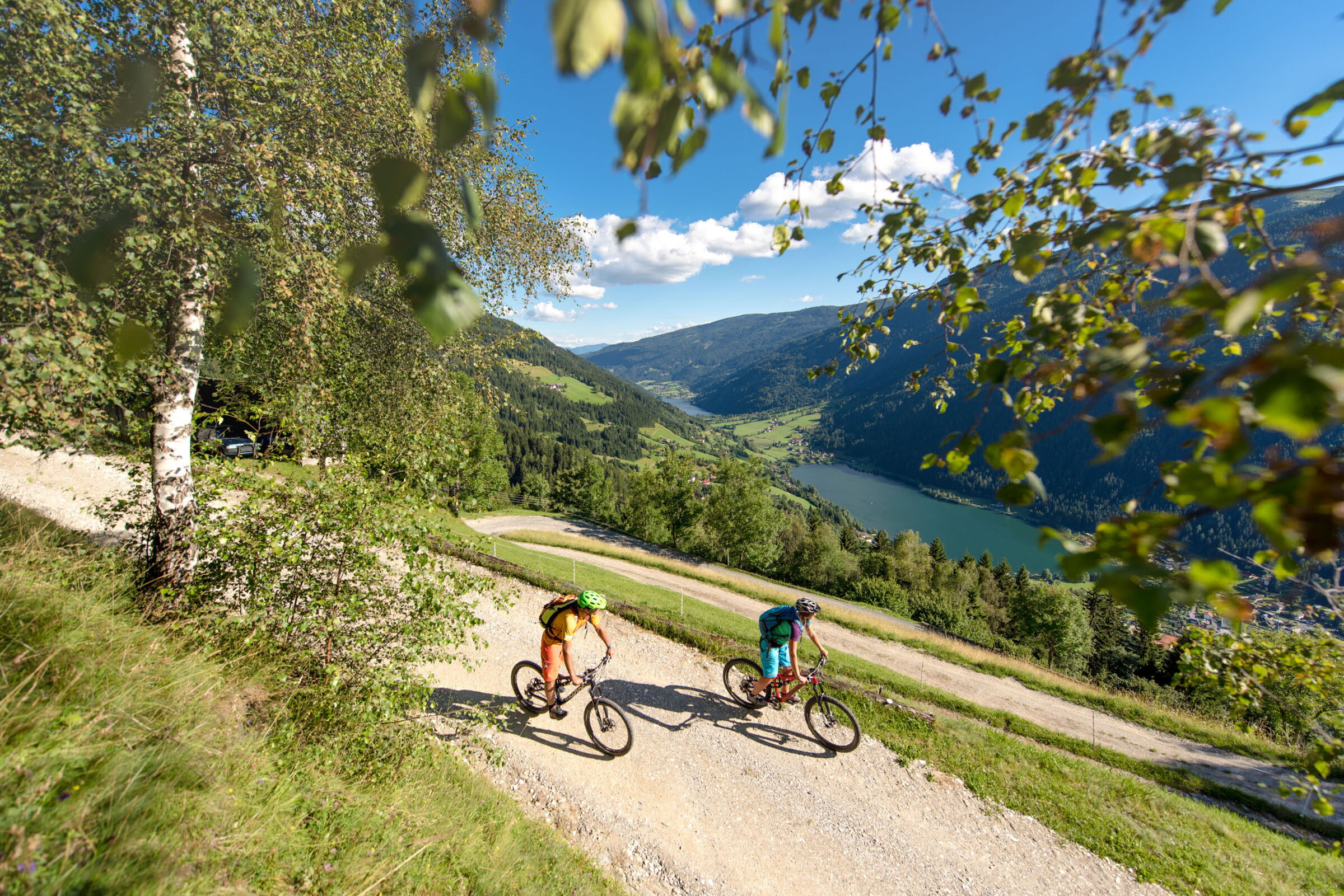 Zwei Radfahrer auf einem Bergweg mit malerischem Talblick und blauem Himmel.